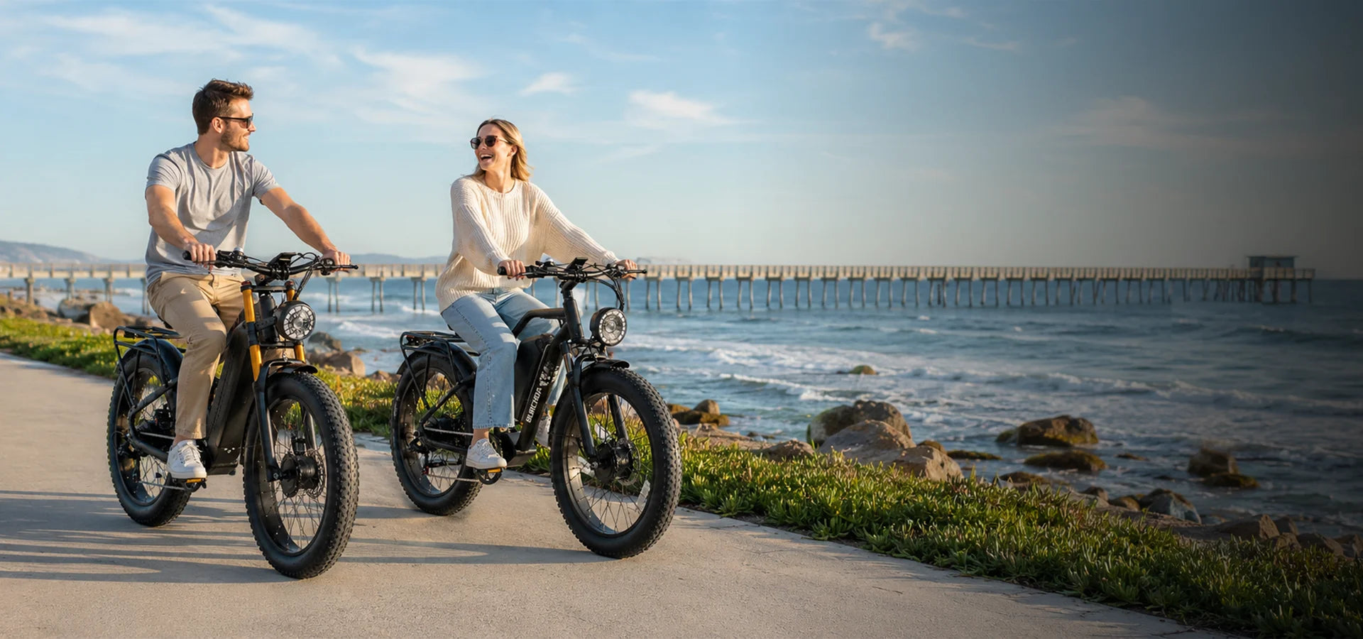 A happy couple riding Burchda AWD fat tire electric bikes along a sunny beach boardwalk. Shop our Summer Sale to upgrade your high-performance beach cruisers and all-terrain ebikes.1621243260e1af0c20-1