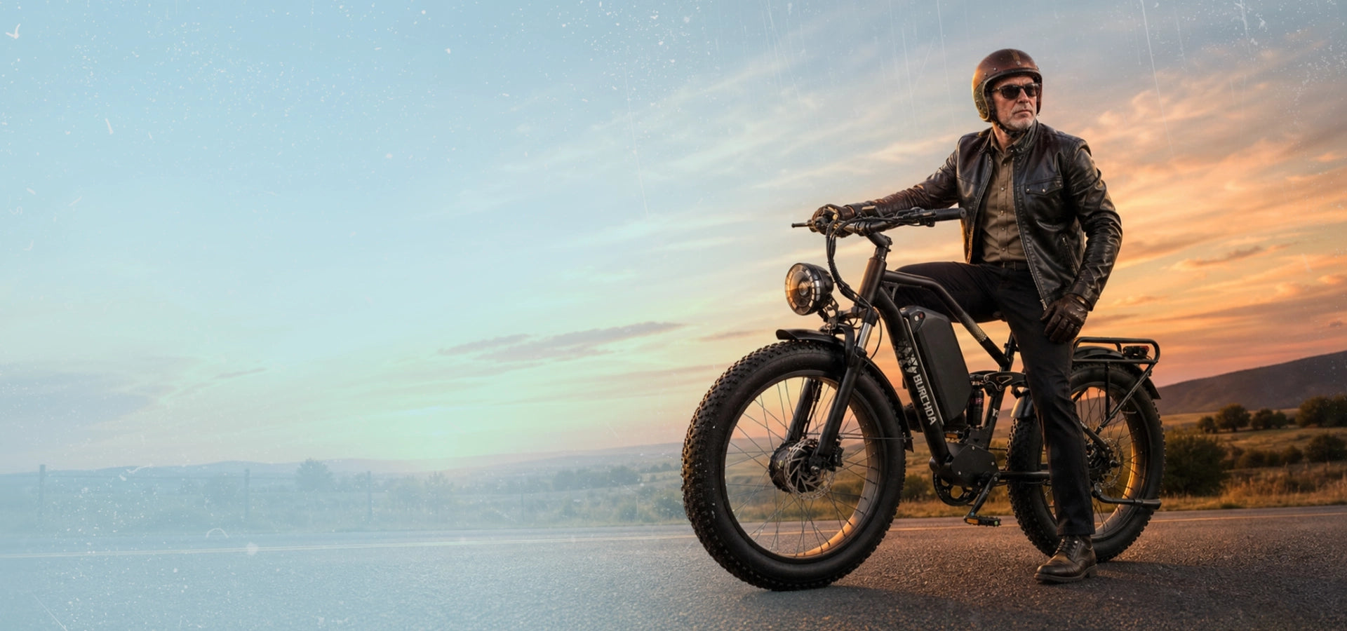 A rider standing over a black Burchda Y3 AWD fat tire electric bike on an open road at sunsetslider_item_Cj6nrE